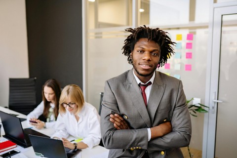 Black man in suit in the foreground with coworkers on laptops at desk in background