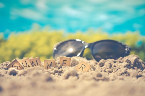 Scrabble tiles spelling 'summer' in sand with sunglasses behind them