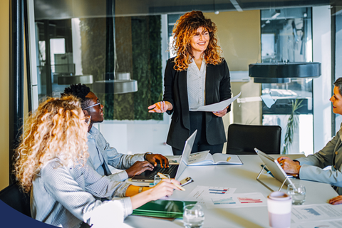 several woman sit around a table while one presents from a paper