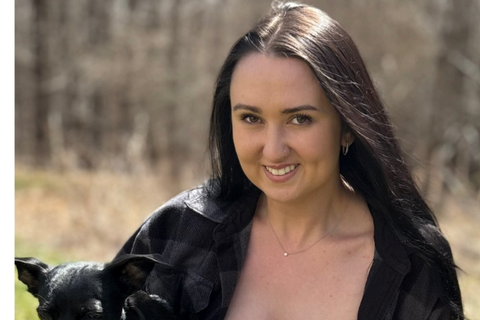 Alumnus wearing an all-black outfit, standing on grass, while holding and embracing a small black dog.