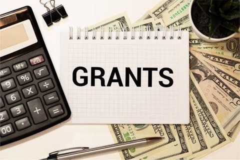 Grants in capital at center of page. On the left is a calculator. On the background is a spread of dollars. On the top right corner is a coffee mug.