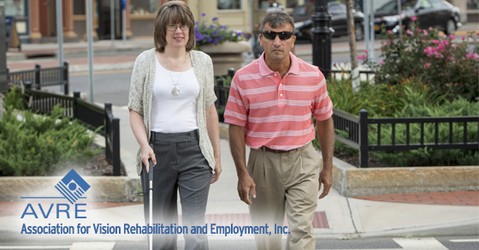 A woman with short brown hair and glasses, wearing a white top, beige cardigan, and dark trousers, holds a white mobility cane while walking alongside a man wearing sunglasses, a pink-and-white striped polo shirt, and khaki pants. They are walking together on a brick-paved urban sidewalk with flowers and parked cars in the background. In the bottom left corner, the logo reads