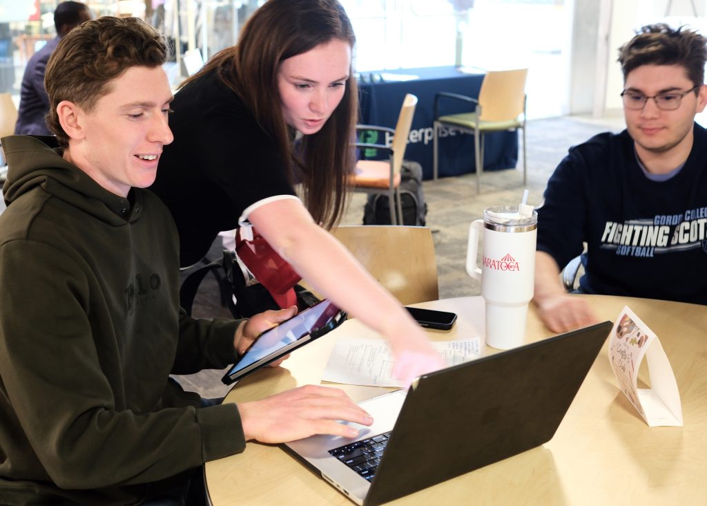 Student seated at computer, woman is standing next to him pointing at screen, another student sits on the right side of the table
