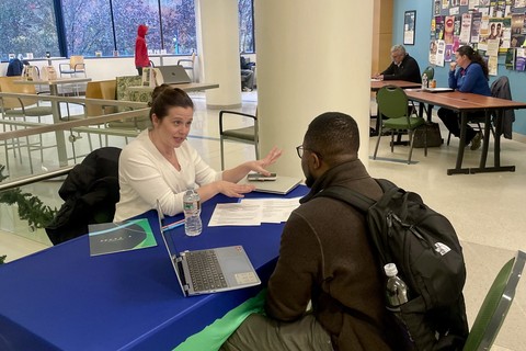 A woman gestures while speaking, across a table from her, a man with a laptop in front of him, leans in.