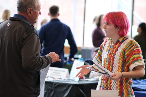 Man in a black jacket stands across from a person with pink hair and a colorful top. The person with pink hair holds some papers and is gesturing with their right hand.