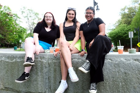3 women, wearing casual clothing, sitting on a stone wall and smiling at the camera.