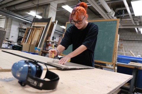Woman with red and pink hair draws with a pencil on a board in a workshop.