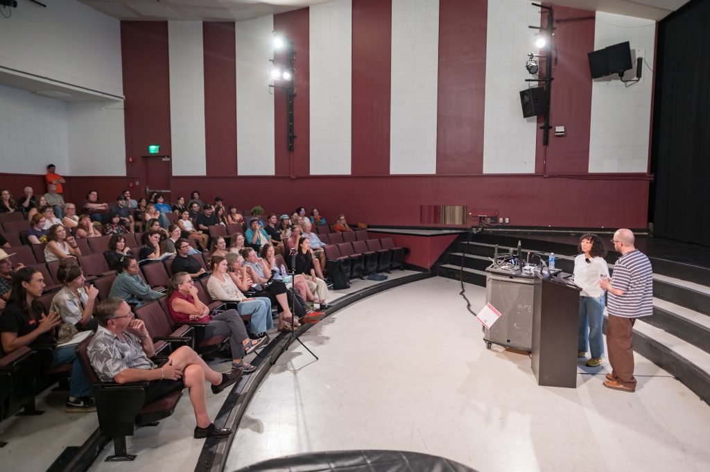 Two professors standing behind a podium while presenting to a crowd inside an auditorium.