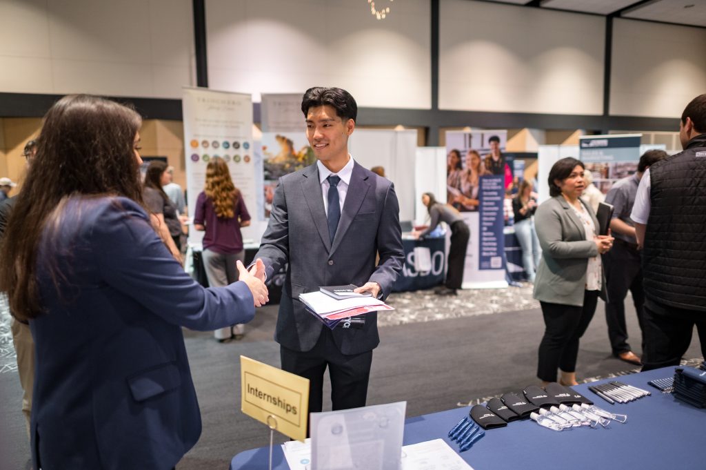 A student wearing a professional suit shakes the hand of an employer during a job fair.