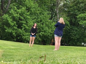 Woman stands in a field posing against a backdrop of trees while another woman takes her picture.