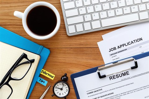 A wooden desk with job application materials, including a printed resume on a clipboard and a job application form. A white keyboard, a cup of black coffee, a blue notebook with sticky notes labeled “Interview” and “Tips,” a pair of black-framed eyeglasses, a silver pen, and a small clock are also present. The scene suggests job search preparation.
