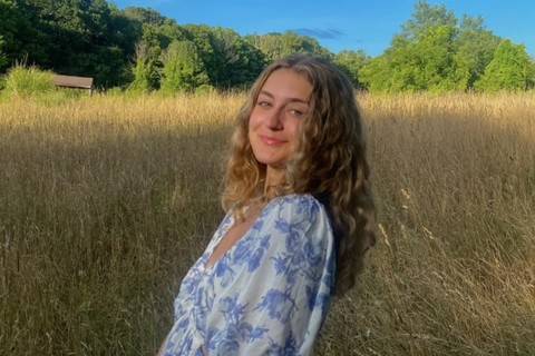 A young woman with wavy, light brown hair stands in a sunlit field of tall, golden grass. She is wearing a white and blue floral dress with short puffed sleeves, and she has a gentle smile on her face. The background features lush green trees under a bright blue sky.