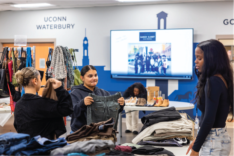 This image depicts the career closet at UConn Waterbury. Several individuals are engaged in sorting and distributing clothing items. In the foreground, a young woman in a hoodie holds up a pair of jeans, while another person next to her examines a piece of clothing. A third woman in a black top and jeans stands nearby. The background features a table with shoes and other donated goods, as well as a large screen displaying a slide with a community outreach theme.
