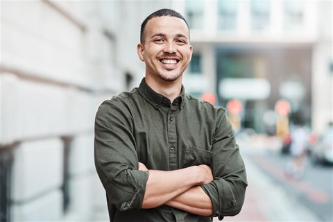 A man smiling confidently with arms crossed, standing on a city street in front of a blurred urban background. He is wearing a dark green button-up shirt and appears relaxed and approachable.