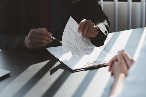 A person in a dark suit holding and pointing to a resume during a job interview, with another person sitting across the table, hands clasped. The scene is lit with natural light casting shadows from window blinds.