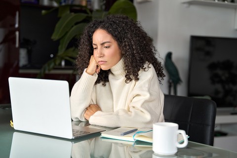 Someone sitting at a table looking at a laptop. Their cheek is resting on one hand, giving the appearance of boredom or frustration.