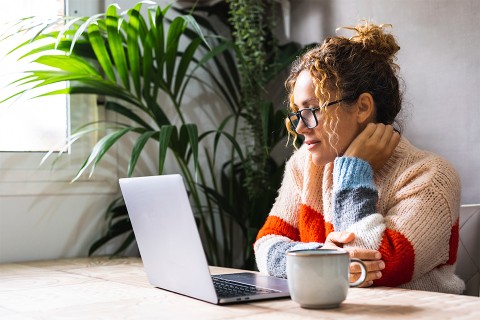 A woman wearing glasses and a colorful sweater sits at a table with a laptop and a mug, looking thoughtfully at the screen.