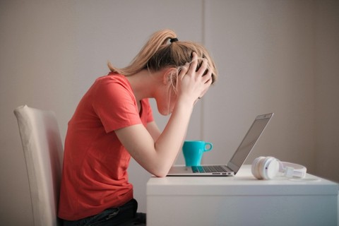 An individual sitting a desk with a laptop and their head in their hands. They appear stressed.