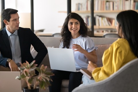 Three people are sitting on a sofa. The person in the middle has a laptop and is speaking to the other two.