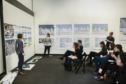 A group of students sit in folding chairs in white walled room. Drawings hang on the wall, and others are on the floor in front of the walls. A professor stands at the front of the room. criticing the art. The artist, a young woman ina grey sweater, stands next to the art, listening to the critique and smiling.