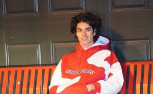 A young man, Richard, wearing a red and white hoodie, sits on an orange bench against a black paneled wall.