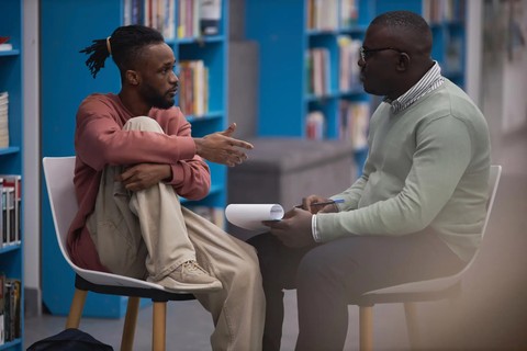 Image shows a male, black student talking to a male, black mental health therapist in a library.