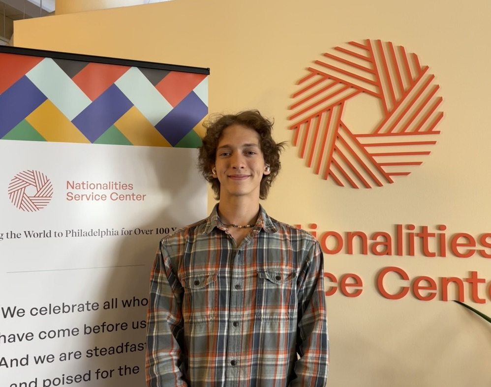 Edwin smiling in a orange and brown plaid shirt in front of the Nationalities Service Center Sign