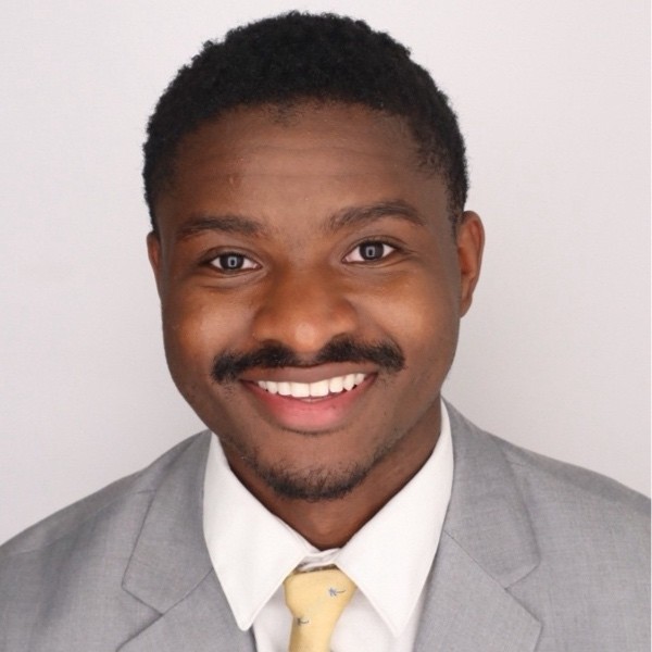 Olakanmi smiling in a grey blazer and yellow tie in front of a white background