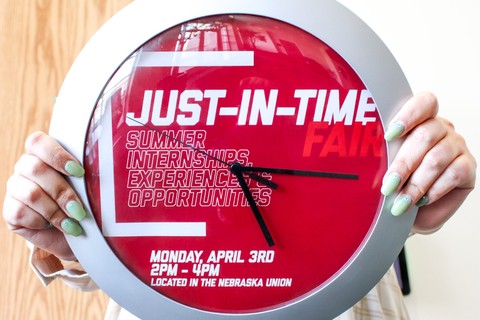 Hands holding a clock with Just-In-Time Fair title and event info listed on the clock face.