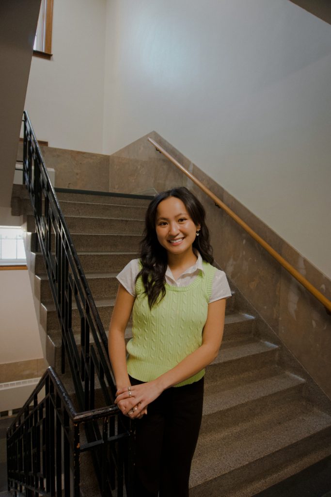 UNL student Sydney Huynh is pictured posing in front of a granite staircase in the Nebraska City Campus Union. Sydney is wearing a light green sweater vest and black dress pants.
