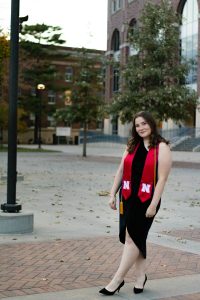 Recent UNL grad, Hailey Abler, pictured outside in a black dress, black heels, and a red graduation ribbon.