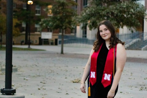 A recent UNL grad, Hailey Abler, pictured outside in a black dress, black heels, and a scarlet UNL graduation ribbon.