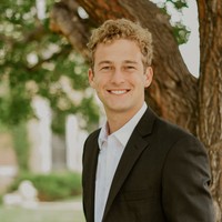 A headshot of Cap Kelly, our Undergraduate Student Employee of the Year, in front of a tree. He is a white man with curly blond hair, wearing a suit.